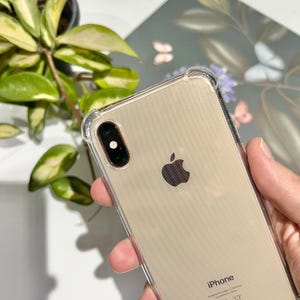 May include: A clear protective phone case on a gold iPhone. The phone has a black camera lens and the Apple logo. The case has reinforced corners. The word "iPhone" is visible at the bottom of the phone.