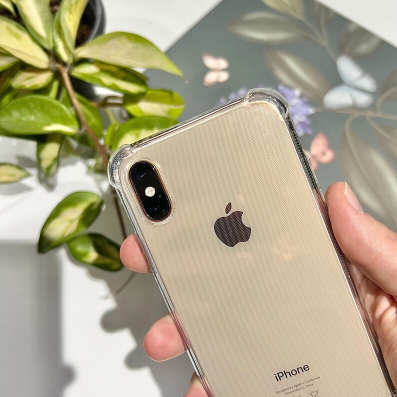 May include: A clear protective phone case on a gold iPhone. The phone has a black camera lens and the Apple logo. The case has reinforced corners. The word "iPhone" is visible at the bottom of the phone.