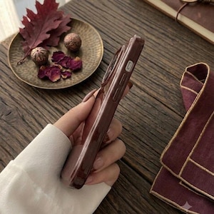 May include: A dark brown smartphone case with a smooth, glossy finish, featuring precise cutouts for side buttons and volume controls. The case is held in a hand, showcasing its slim profile and deep, solid color against a dark wooden surface.