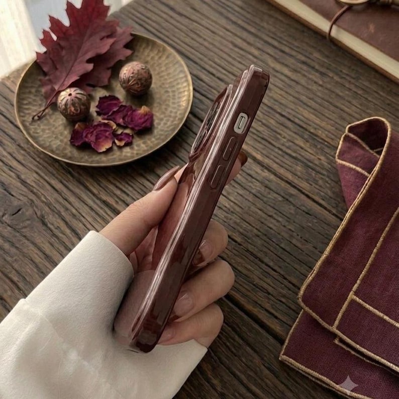 May include: A dark brown smartphone case with a smooth, glossy finish, featuring precise cutouts for side buttons and volume controls. The case is held in a hand, showcasing its slim profile and deep, solid color against a dark wooden surface.