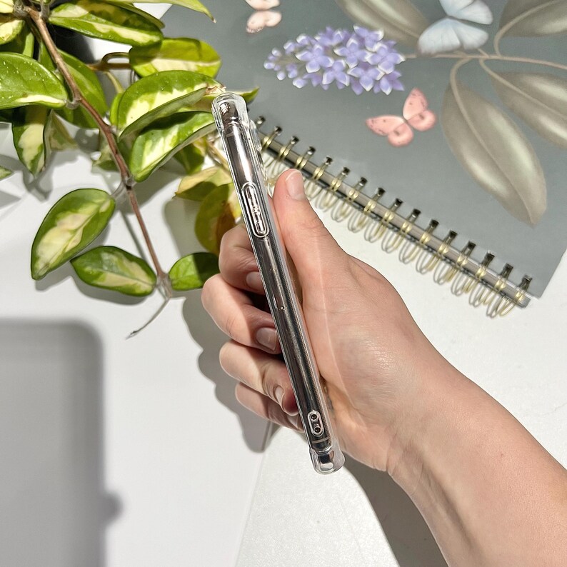 May include: Clear phone case held in a hand. The case is made of transparent plastic and has a slim design. The phone case is held near a spiral-bound notebook with a floral design and a green plant.