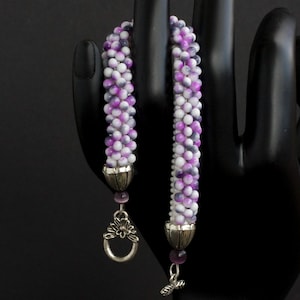 Può includere: Un braccialetto di perline con un motivo a spirale di perline bianche, viola e grigie. Il braccialetto ha terminali argentati e una chiusura decorativa con un motivo floreale. Un piccolo ciondolo pende dalla chiusura.