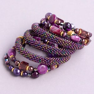 Può includere: Una collezione di braccialetti di perline viola. I braccialetti presentano una varietà di perline, tra cui forme rotonde, quadrate e irregolari, in tonalità di viola, oro e colori iridescenti. I braccialetti sono impilati, creando un effetto a strati.