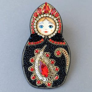 Op de afbeelding: Een handgemaakte broche in de vorm van een Matroesjka-pop. De pop heeft een zwart, met kralen bezet lichaam, een geschilderd gezicht en is versierd met rode en gouden versieringen. De broche is ongeveer 7.6 cm hoog.