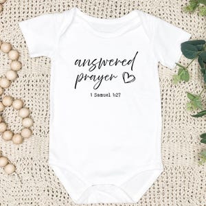 May include: White baby onesie with black text that reads "answered prayer" with a small heart symbol and the scripture reference "1 Samuel 1:27".