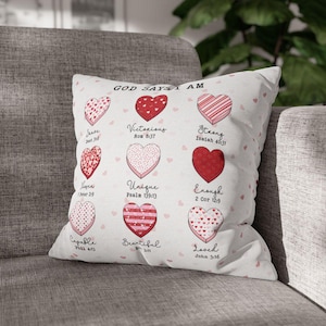 Puede incluir: Un cojín decorativo con fondo blanco y un diseño de corazones rosas y rojos. El cojín presenta el texto "GOD SAYS I AM" y varias afirmaciones como "Victorious" y "Loved", cada una acompañada de un versículo bíblico.