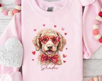 May include: Pale pink sweatshirt with a watercolour illustration of a dog wearing heart-shaped glasses and a polka-dot bow tie. The dog's name, "Shadow," is written in cursive below. Red hearts are scattered around the dog.
