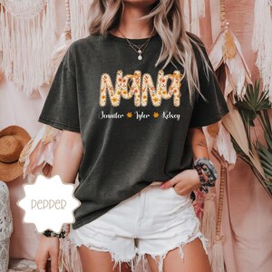 May include: Dark gray t-shirt with the word "NANA" in a floral design. Below the word "NANA" are the names "Jennifer", "Tyler", and "Kelsey". The shirt is a comfortable, casual style.