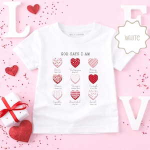 Op de afbeelding: Wit Bella Canvas T-shirt met de tekst "GOD SAYS I AM" en hartafbeeldingen. Elk hart heeft een woord als "Brave" of "Loved" en een bijbelvers. Het shirt is omgeven door Valentijnsdagdecoraties.