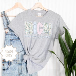 May include: Sport grey t-shirt featuring the word "NICU" in a floral and gingham design, with the text "little patients, big futures" and the Bible verse Jeremiah 29:11. The shirt is displayed on a wooden hanger.