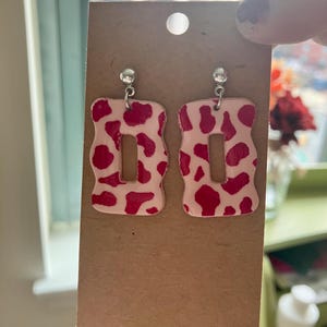 May include: A pair of pink and white cow print earrings with a silver hook. The earrings are shaped like a rectangle with a cutout in the middle.