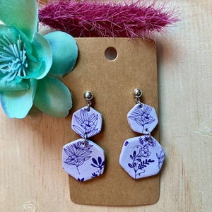 May include: A pair of lavender dangle earrings with a floral pattern in blue. The earrings are made of polymer clay and have a silver hook.