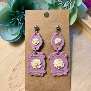 May include: A pair of lavender-colored polymer clay earrings with white flower accents. Each earring features a double-layered design with a smaller flower charm on top and a larger flower charm on the bottom.