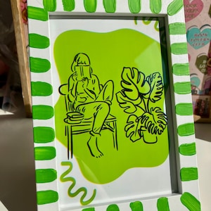 May include: A framed art print featuring a black line drawing of a person reading, a potted plant, and a lime green background. The white frame is decorated with green brushstrokes.