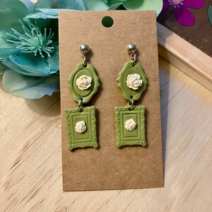 May include: A pair of green and white floral dangle earrings. The earrings feature a small white flower in a green frame, followed by a larger white flower in a larger green frame.