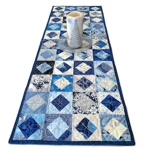 May include: A blue and white quilted table runner with a diamond pattern. The runner is decorated with a silver snowflake design. A white candle is placed on top of the runner.