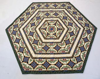 May include: Hexagonal quilted table topper with a green border. The center features a cream-colored background with blue and green floral patterns. The design includes red ribbons and a repeating geometric pattern.