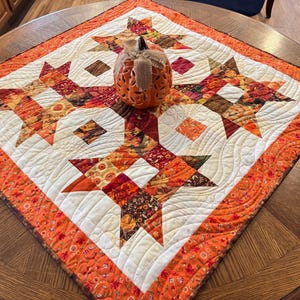 May include: A square fabric quilt featuring a star pattern in shades of orange, red, brown, and cream, with intricate swirling machine quilting. An orange ceramic pumpkin with cut-out details and a burlap ribbon sits in the center of the quilt.