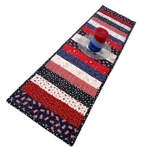 May include: A patriotic table runner with red, white, and blue stripes and star patterns. A red, white, and blue candle sits on a clear glass plate. The runner is quilted and has a dark blue border.