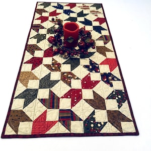 May include: A rectangular quilted table runner featuring a patchwork star pattern in shades of red, navy blue, brown, and cream. The runner is bordered in dark red and topped with a red pillar candle surrounded by a decorative berry wreath.
