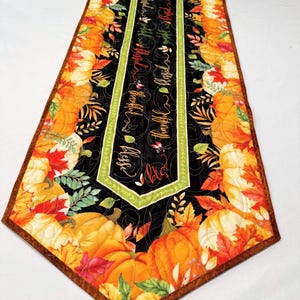 May include: A quilted table runner with a fall theme. The runner features a black center panel with the words "bless", "hope", "thankful", and "love" in a script font. The edges are decorated with pumpkins, leaves, and a brown border.