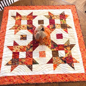 May include: A square fabric quilt featuring a star pattern in shades of orange, red, brown, and cream, with wavy quilting lines. Centered on the quilt is a hollow orange ceramic pumpkin decoration adorned with a tan burlap ribbon bow on top.
