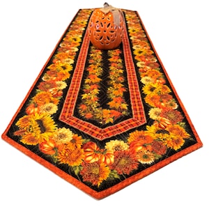 May include: A quilted table runner featuring a black background with a vibrant print of sunflowers, orange pumpkins, and autumn leaves, accented by red and orange plaid borders, topped with a decorative orange ceramic pumpkin figurine.