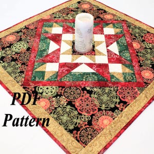 Puede incluir: Camino de mesa acolchado cuadrado con un diseño de estrella central. El camino de mesa presenta patrones geométricos rojos, verdes, dorados y blancos, con un borde floral negro y un ribete dorado. Una vela blanca con un diseño de copo de nieve se encuentra en el centro. El texto "PDF Pattern" es visible.