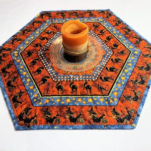 May include: Hexagonal Halloween-themed table runner with orange, blue, and yellow accents. The fabric features black cat and witch silhouettes, along with star patterns. A lit orange candle sits on a decorative glass plate in the center.