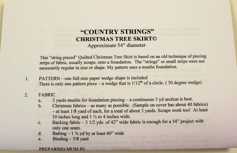 以下が含まれることがあります： 「COUNTRY STRINGS」クリスマスツリースカートの説明が印刷された紙。直径約137cm。テキストには、モスリン、クリスマス生地、裏地、中綿、バインディングなど、生地の要件が詳しく記載されています。