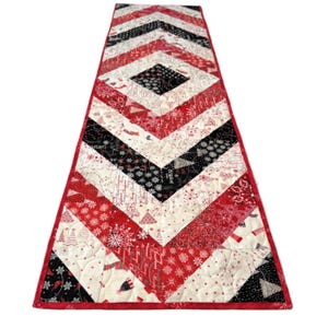 May include: A quilted Christmas table runner with a red border. The runner features a chevron pattern in red, black, and white fabrics with holiday-themed designs, including snowflakes and trees. The center has a diamond-shaped design.