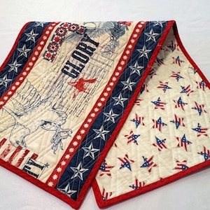 May include: A quilted table runner with a patriotic theme. The runner features a cream-colored background with a large eagle illustration, the word "GLORY", and red, white, and blue accents. The reverse side has a pattern of red, white, and blue stars.