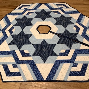 May include: An octagonal fabric tree skirt featuring a geometric star pattern in shades of navy blue, light blue, and white. The quilted textile has a central hexagonal opening with a straight slit for placement around a tree base.