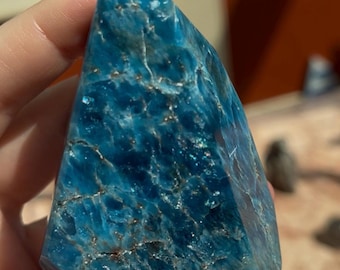 May include: A faceted, triangular-shaped blue apatite crystal. The crystal has a deep, saturated blue color with lighter blue and white veining. The surface has a slightly rough texture with some areas of sparkle. The crystal is held in a hand.
