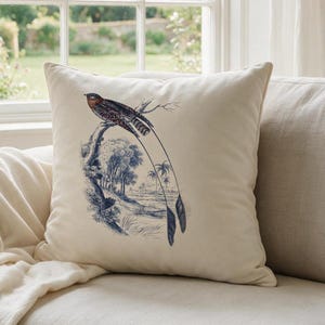 Op de afbeelding: Een vierkant gebroken wit sierkussen met een gedetailleerde blauwe en bruine illustratie van een vogel met een lange staart op een tak boven een landschap met bomen en gebladerte. Het kussen is gemaakt van gladde stof en heeft een effen achterkant.