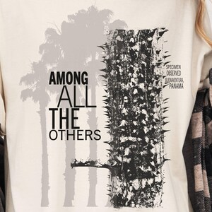 May include: A cream-colored t-shirt featuring a graphic print with the text AMONG ALL THE OTHERS, a silhouette of palm trees, and a textured black and white image of a tree trunk with the text SPECIMEN OBSERVED BUENAVENTURA PANAMA.