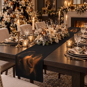 Puede incluir: Un camino de mesa negro con una ilustración dorada y etérea de un ángel, sobre una mesa de comedor de madera oscura preparada para una comida festiva con candelabros dorados, cristalería y un centro de mesa de piñas y flores.
