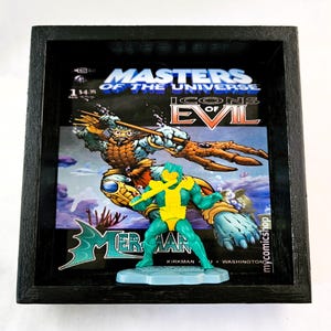 May include: A framed Masters of the Universe comic book featuring the character Mer-Man. The comic book cover features the title "Icons of Evil" and a blue and green illustration of Mer-Man. A plastic action figure of Mer-Man is displayed in front of the comic book.