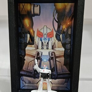 以下が含まれることがあります： 白と黒のおもちゃのロボットのフィギュアが、より大きなロボットのイラストの前に配置された額入りのアートワーク。 おもちゃは白い台座の上に立っており、背景にはオレンジと白のアクセントが施されたロボットの詳細なイラストが描かれています。 フレームは黒です。