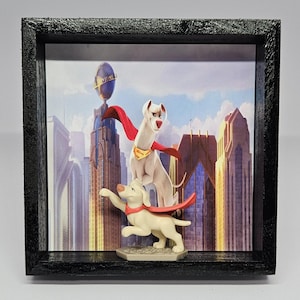 May include: A framed shadow box featuring two dog figurines in superhero attire. The top dog is white with a red cape and gold emblem. The bottom dog is light tan with a red cape. The background depicts a cityscape with a blue sky.