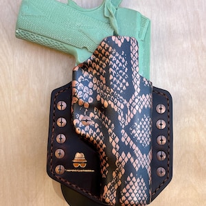 May include: A custom leather holster with a snakeskin pattern in black and copper tones. The holster is secured with copper rivets and features a logo with a hat and sunglasses. A green gun is inside the holster.