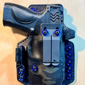 Peut inclure: Un étui en cuir noir avec des détails bleus et un motif camouflage. Il est équipé d'un clip métallique et de vis bleues. La couture est bleue. Conçu pour contenir une arme à feu, l'étui est robuste et durable.