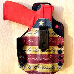 Pu&ograve; includere: Una pistola rossa &egrave; alloggiata in una fondina con un design patriottico della bandiera americana. La fondina presenta accenti rossi, bianchi e blu, con il testo "to the security of Arms, shall not be." La fondina ha una clip nera.