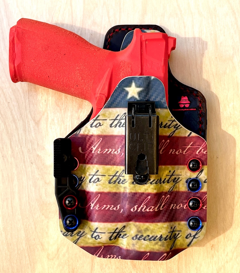 Pu&ograve; includere: Una pistola rossa &egrave; alloggiata in una fondina con un design patriottico della bandiera americana. La fondina presenta accenti rossi, bianchi e blu, con il testo "to the security of Arms, shall not be." La fondina ha una clip nera.