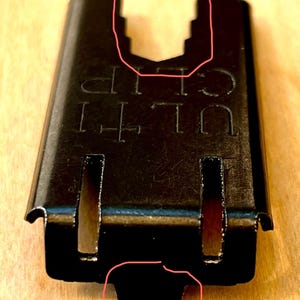 Pu&ograve; includere: Primo piano di una ULTI CLIP in metallo nero. La clip ha una forma rettangolare con una parte superiore dentellata e due fessure verticali. Il testo "ULTI CLIP" &egrave; stampato sulla superficie. La clip &egrave; progettata per fissare oggetti.