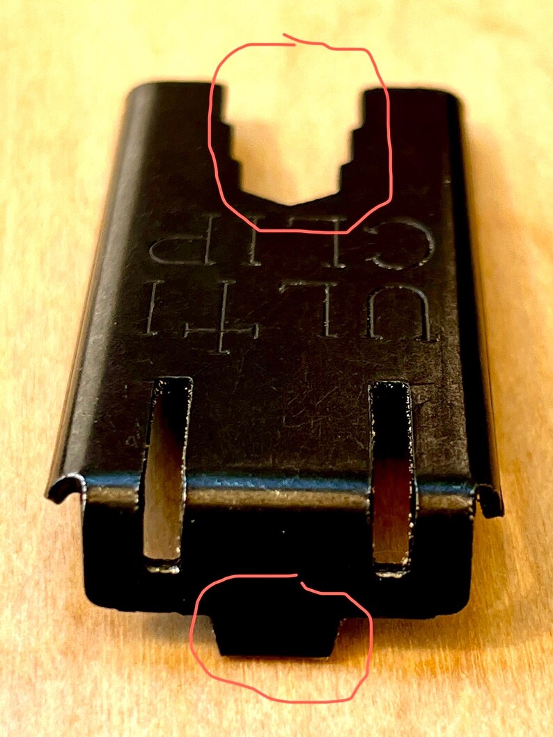 Pu&ograve; includere: Primo piano di una ULTI CLIP in metallo nero. La clip ha una forma rettangolare con una parte superiore dentellata e due fessure verticali. Il testo "ULTI CLIP" &egrave; stampato sulla superficie. La clip &egrave; progettata per fissare oggetti.