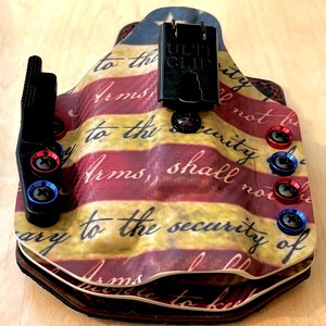 Pu&ograve; includere: Una fondina personalizzata con un design patriottico. La fondina presenta un motivo a strisce rosse e gialle con il testo "Arms, shall not be" e "to the security of". Ha hardware nero e viti rosse e blu.