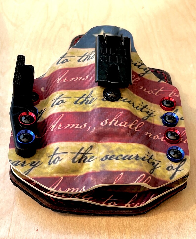 Pu&ograve; includere: Una fondina personalizzata con un design patriottico. La fondina presenta un motivo a strisce rosse e gialle con il testo "Arms, shall not be" e "to the security of". Ha hardware nero e viti rosse e blu.