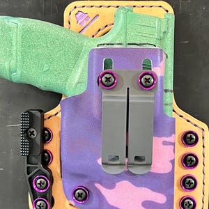 Op de afbeelding: Een paarse en roze camouflage leren holster met een zwarte clip en paarse schroeven. De holster is ontworpen om een ​​vuurwapen te houden.