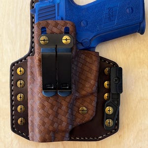 May include: A brown leather holster with a basketweave pattern, holding a blue handgun. The holster has black clips and brass-colored hardware. The stitching is white, and the background is a light wood grain.
